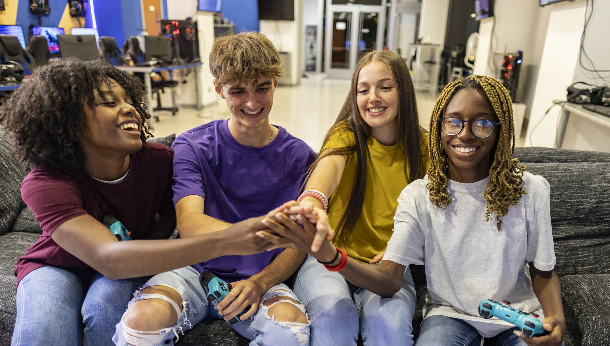 Group of friends joining hands and smiling after playing video games together sitting on a sofa in an internet cafe, they're holding gamepads and having fun