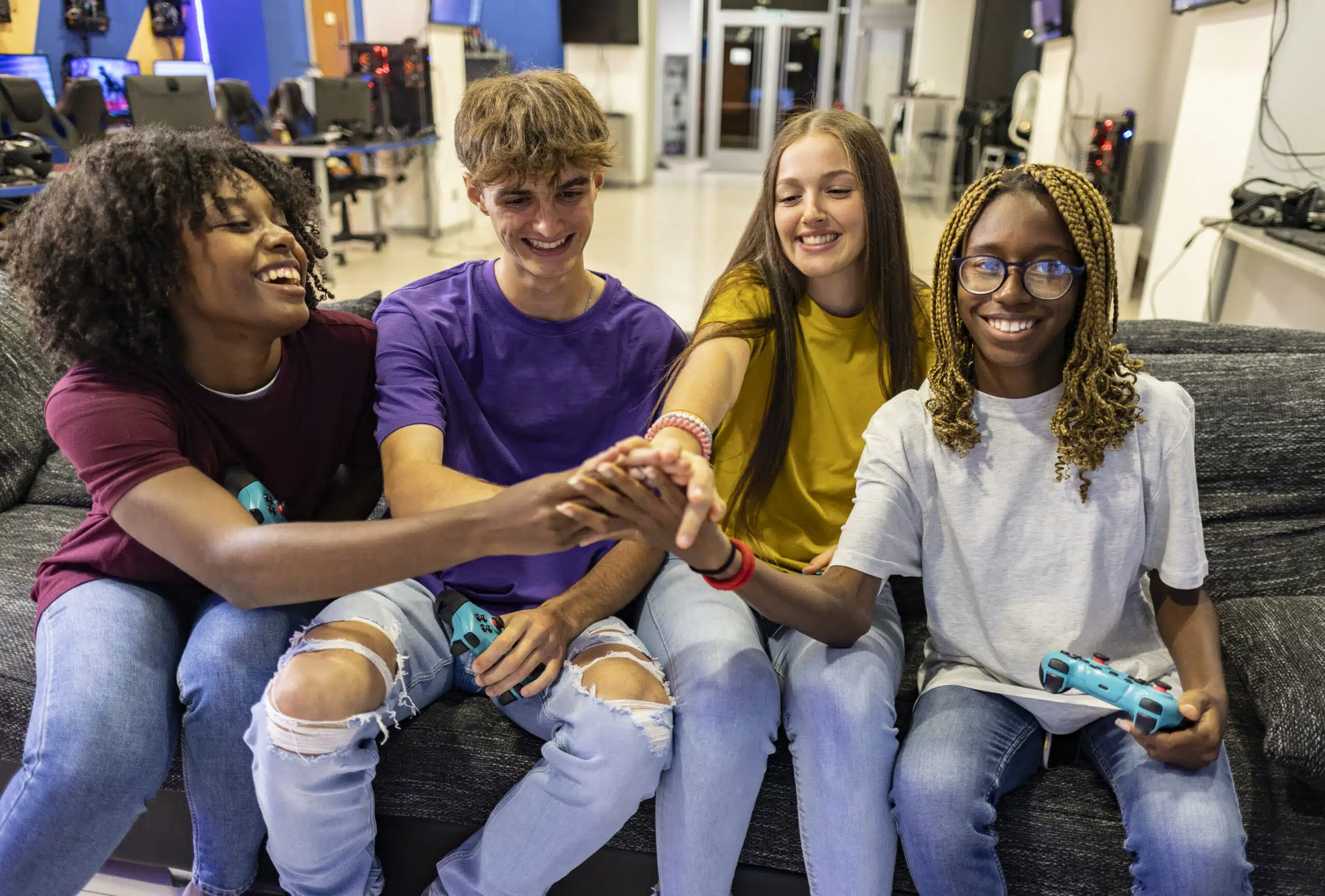 Group of friends joining hands and smiling after playing video games together sitting on a sofa in an internet cafe, they're holding gamepads and having fun