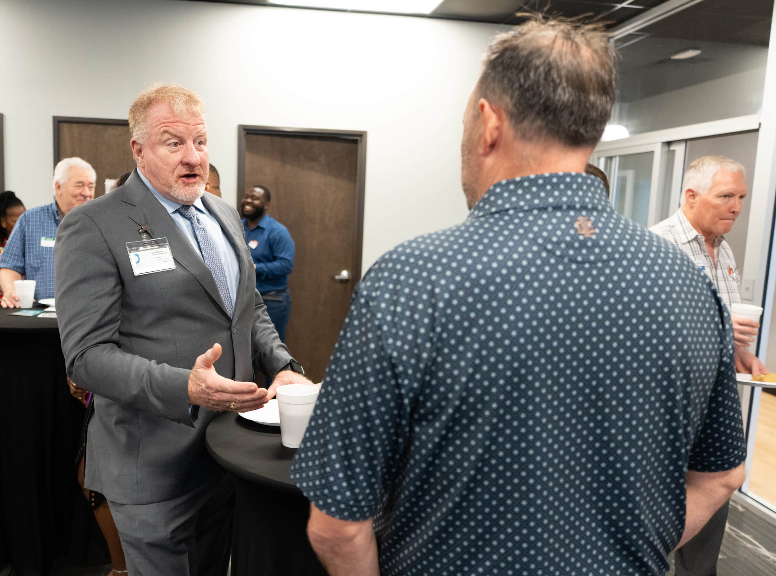 Dan B. Dalton of P3:14 Consulting converses with another attendee while networking at a professional event, wearing a gray suit and name badge.