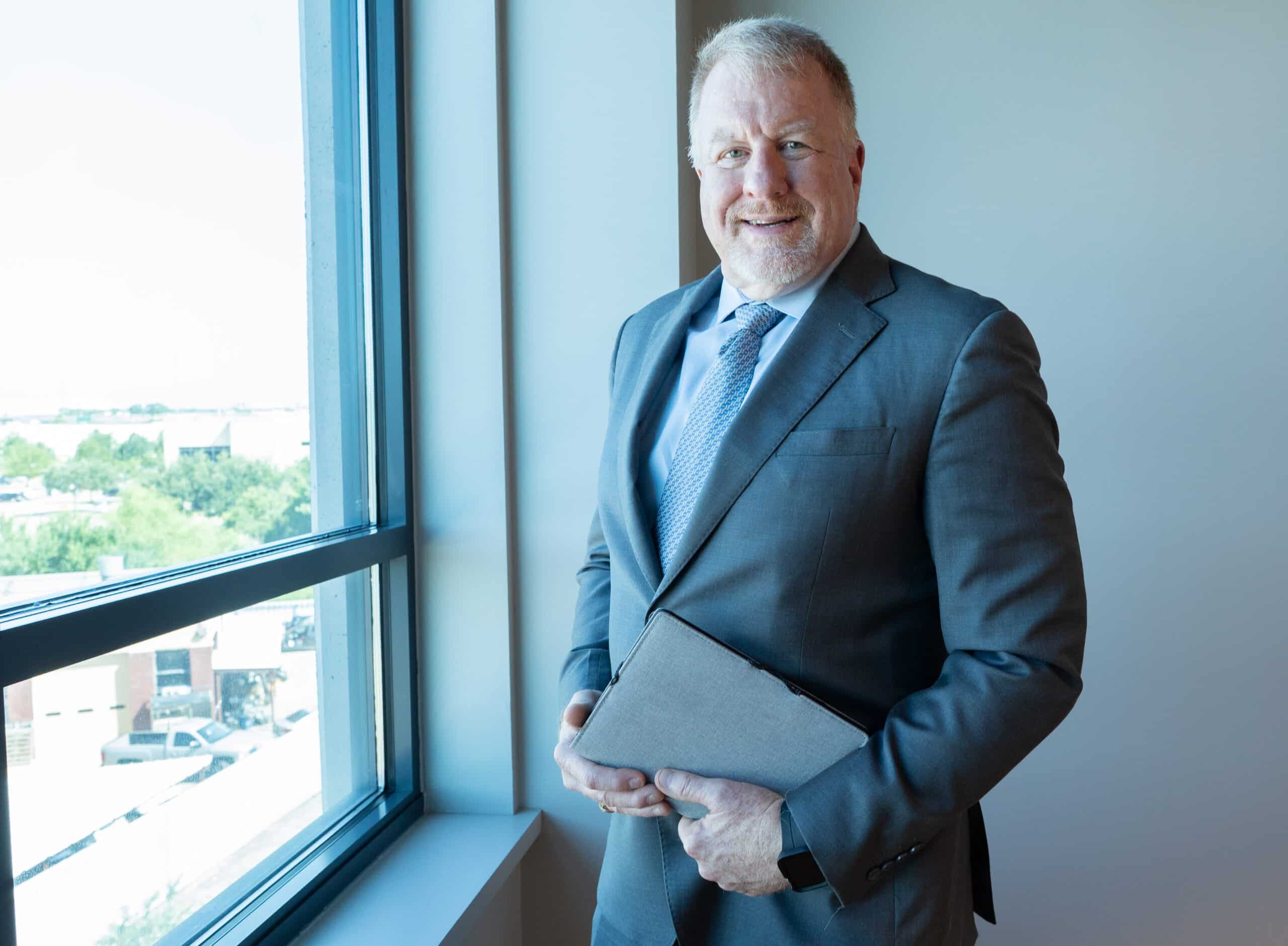 Dan B. Dalton, owner of P3:14 Consulting, stands by a large office window holding a gray tablet, dressed in a professional gray suit with a blue shirt and tie.