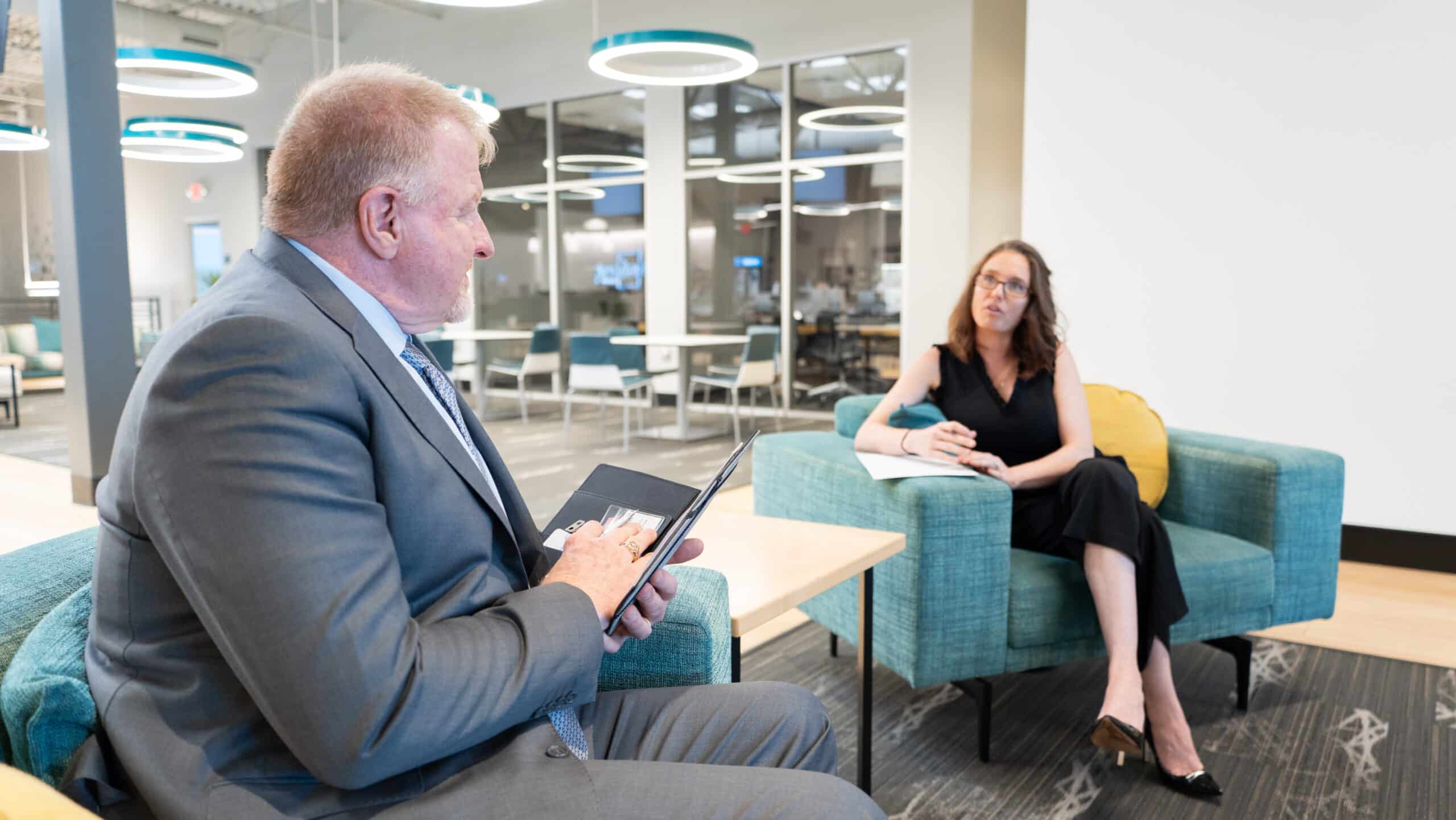 Dan B. Dalton of P3:14 Consulting speaks with a woman taking notes during a one-on-one meeting in a modern office lounge with teal furniture and circular lighting.
