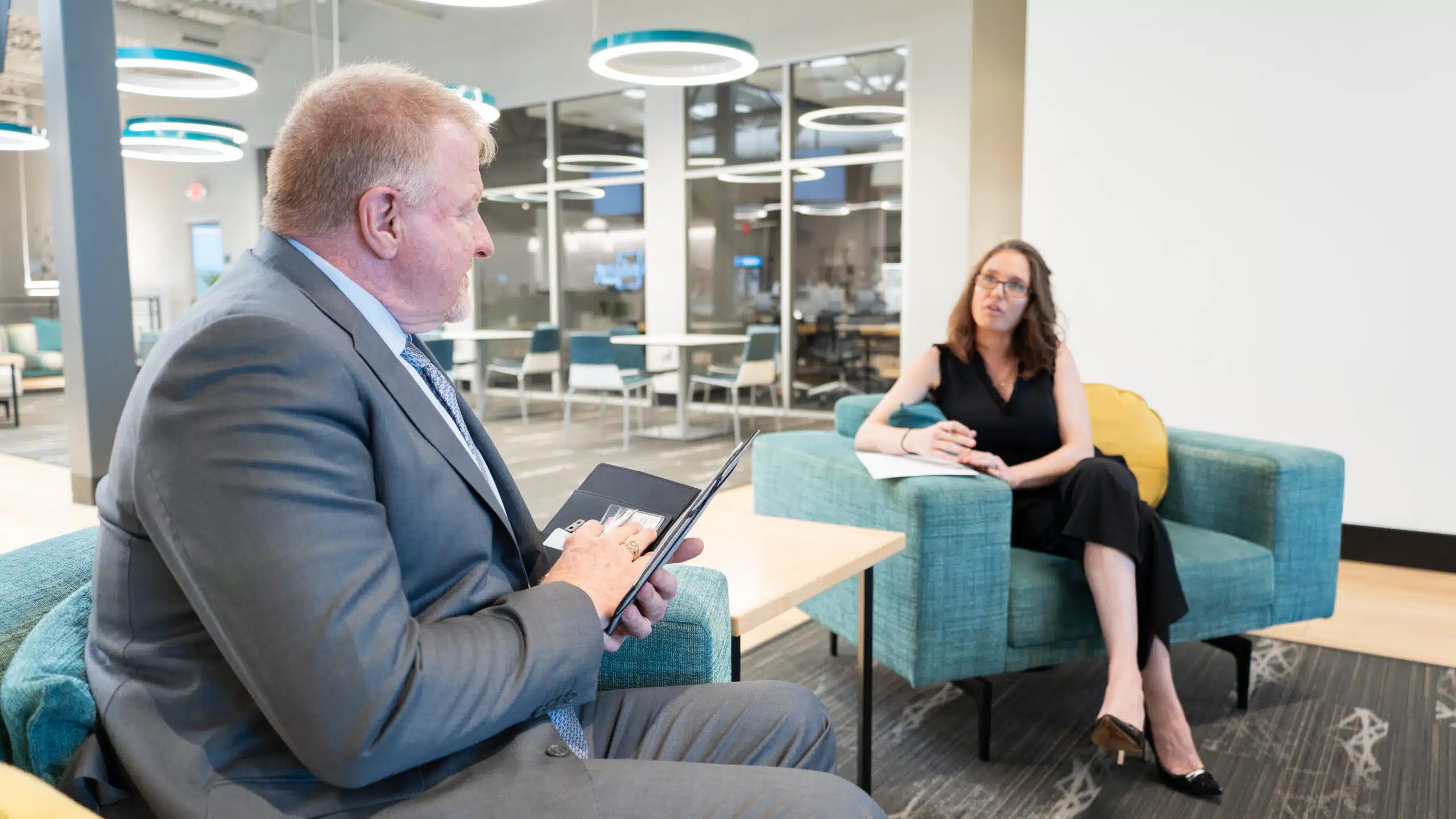 Dan B. Dalton of P3:14 Consulting speaks with a woman taking notes during a one-on-one meeting in a modern office lounge with teal furniture and circular lighting.