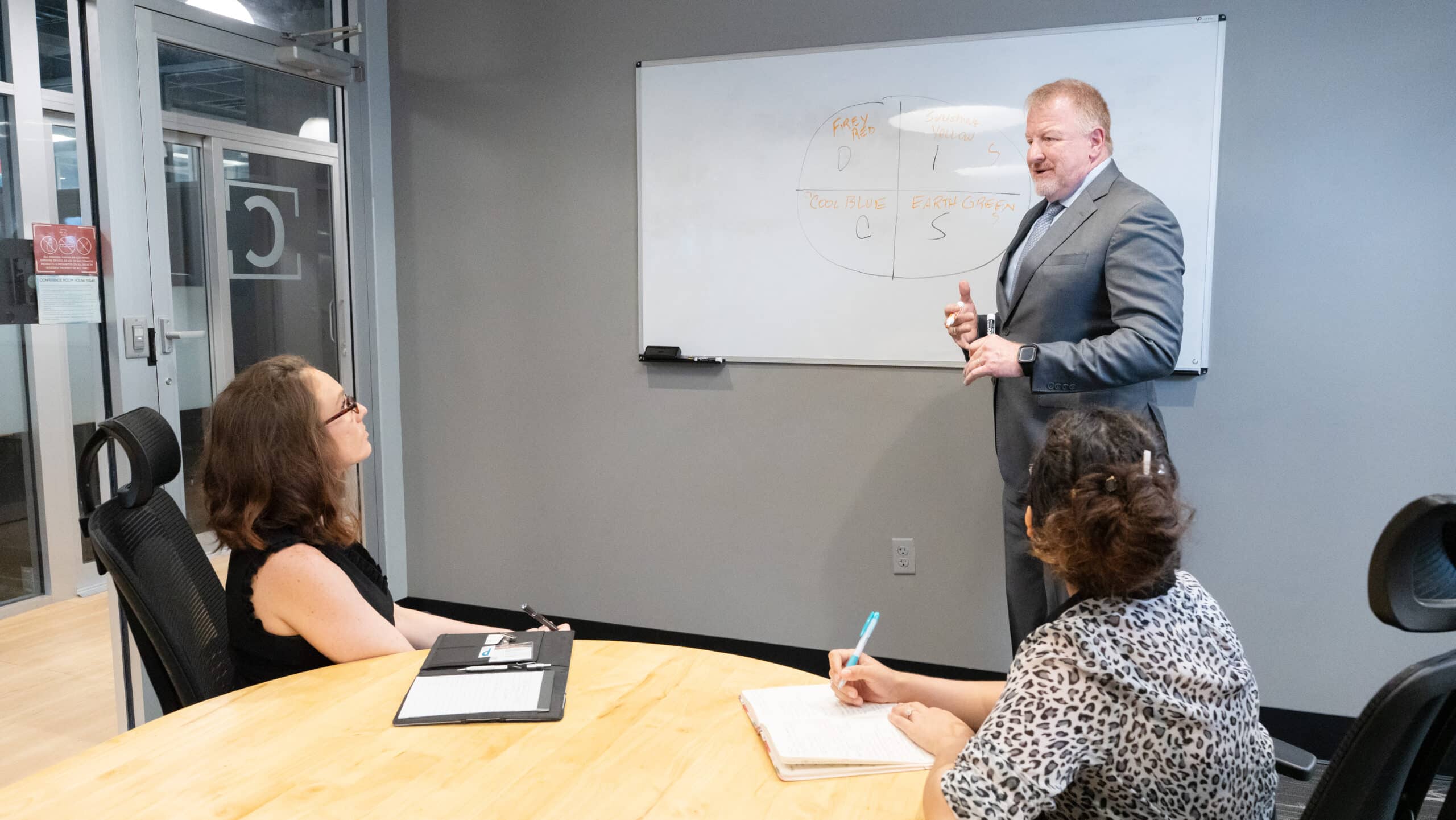 Dan B. Dalton, owner of P3:14 Consulting, gives a presentation using a whiteboard during a strategy meeting with two women seated at a round table in a modern office.